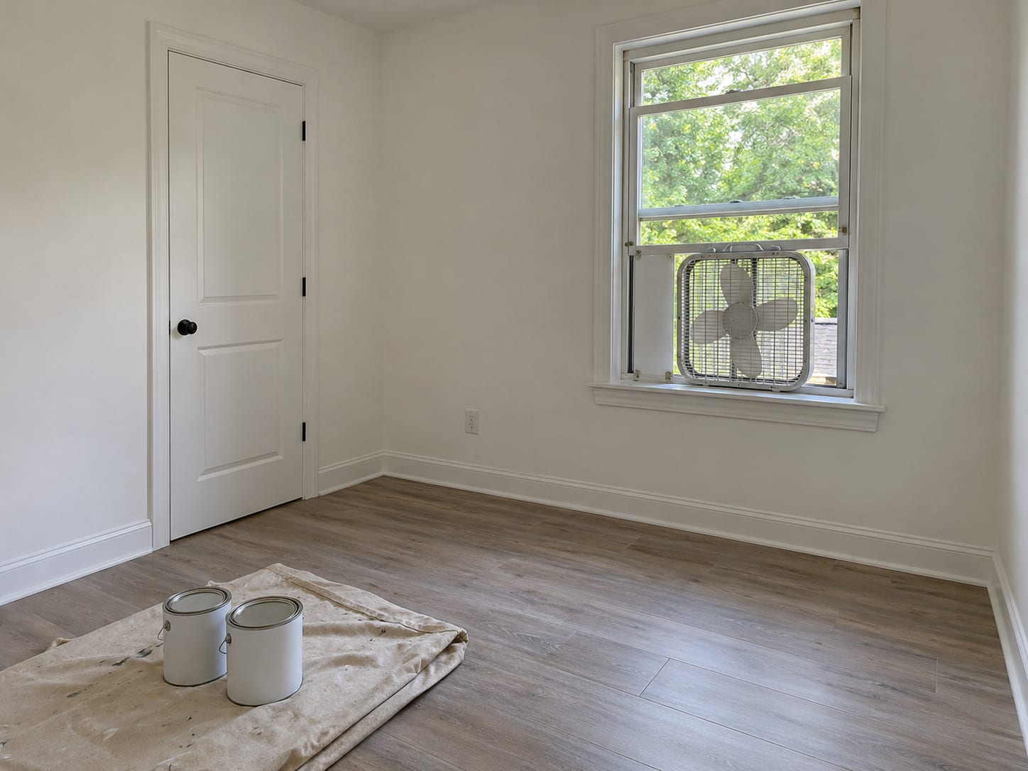 A freshly painted empty bedroom in a New Jersey house with the window open and a fan secured in it, and closed paint cans on a drop cloth