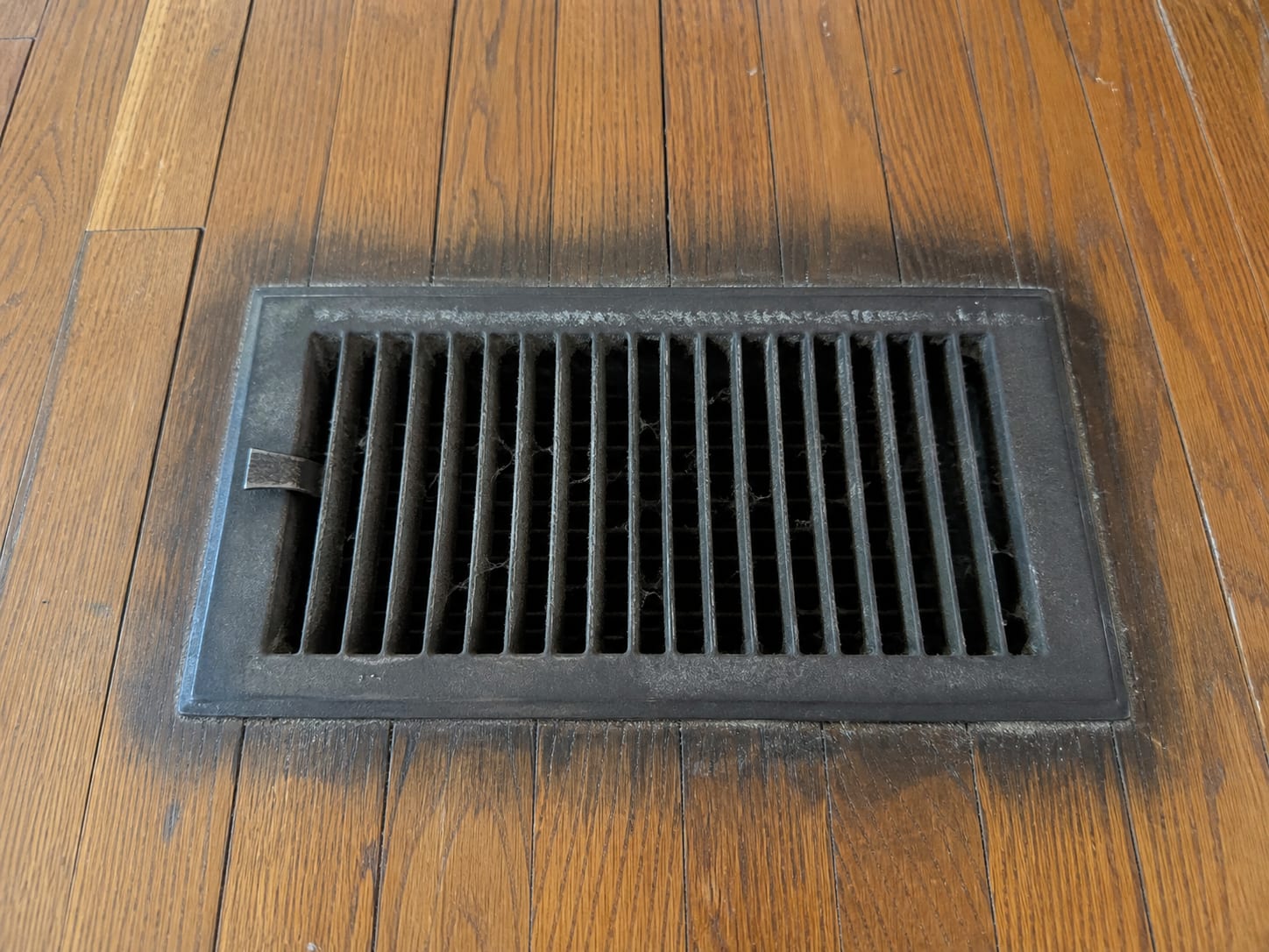 A metal floor supply register set in hardwood with dust furring the vanes and a soft dark halo staining the finish in a ring around the opening, in Alpine, New Jersey