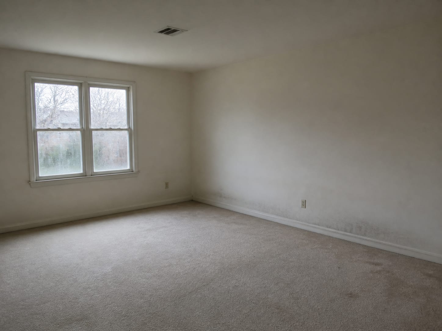 A wider view of an empty room in an East Windsor townhome with a closed window carrying condensation film along the lower glass, a faint dark band low on the exterior wall and a ceiling supply register above
