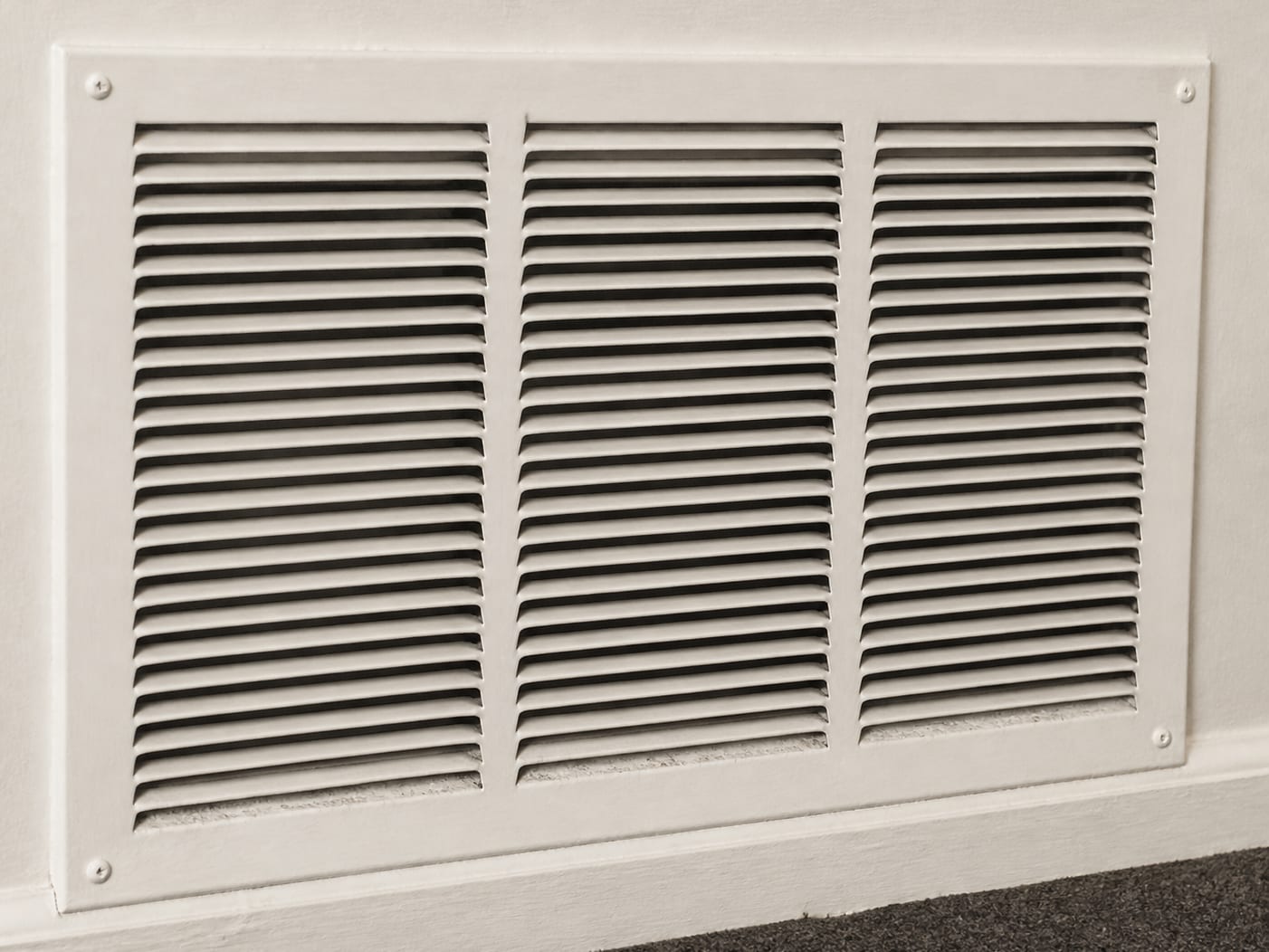 A close view of a painted metal return air grille in a Livingston hallway wall, with fine dust drawn onto the louver edges showing how air moves through the house