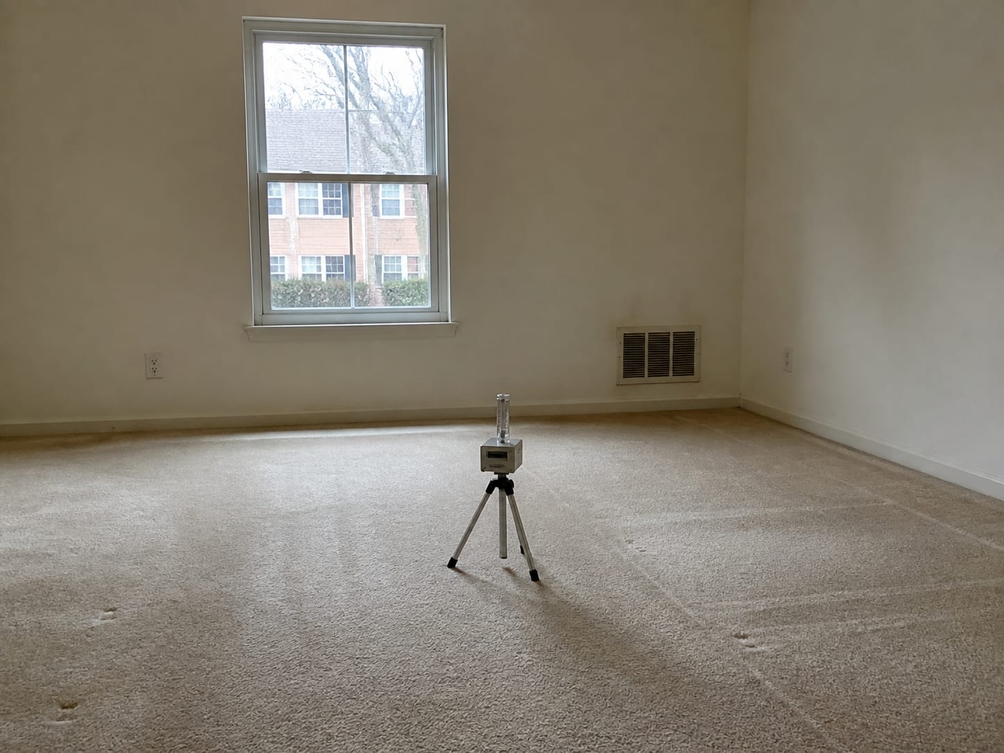 An air sampling pump on a low tripod standing in the middle of an empty carpeted condominium living room, the window shut and latched behind it and a return air grille set into the wall, in Plainsboro, New Jersey