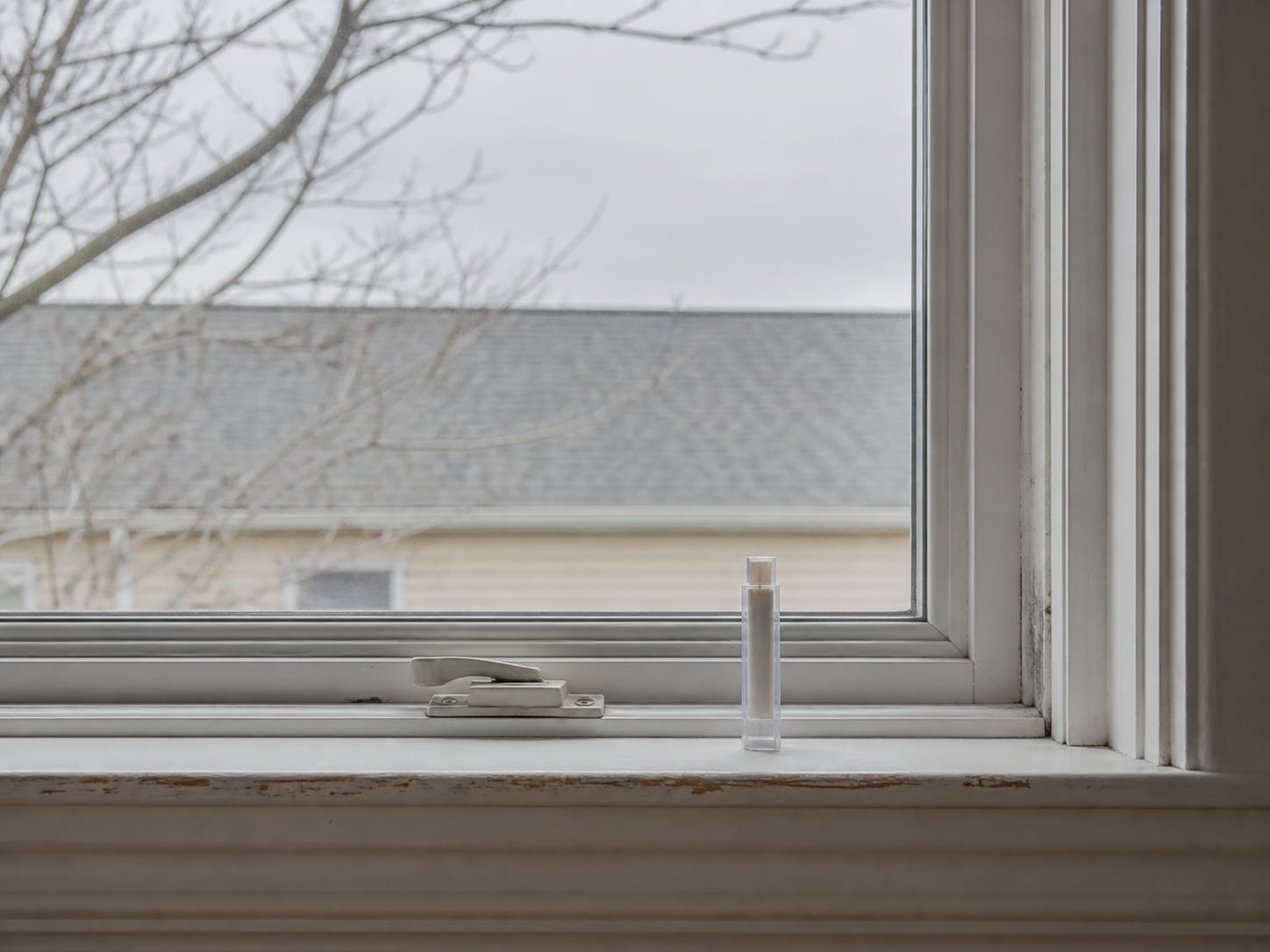A closed and latched window in an empty room with a sealed sample cassette standing upright on the sill beside the catch, bare walls and no furnishings, in Plainsboro, New Jersey