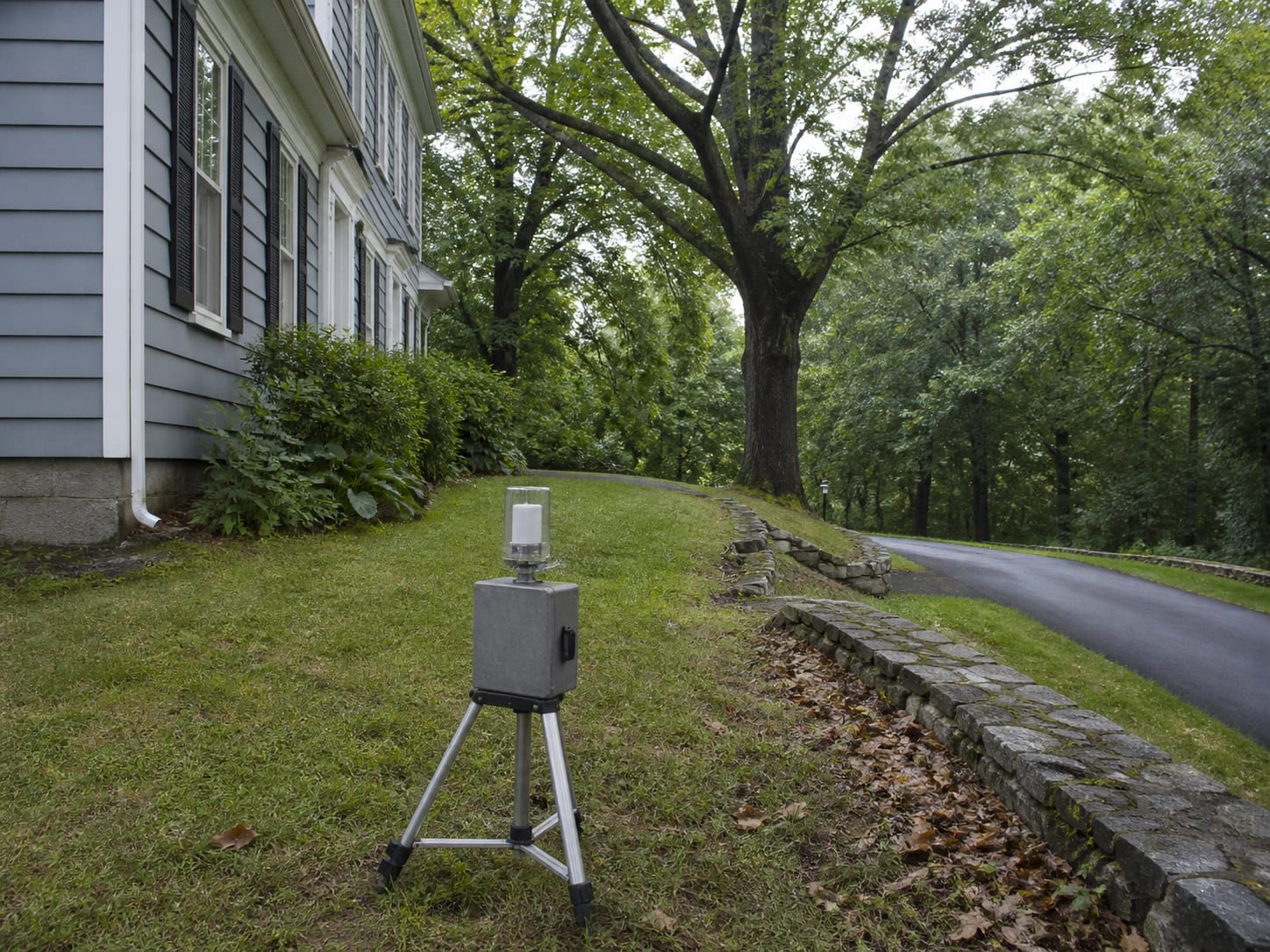 An air sampling pump on a tripod on a lawn beneath mature deciduous trees beside a clapboard house, taking the same day outdoor reference, in Warren, New Jersey