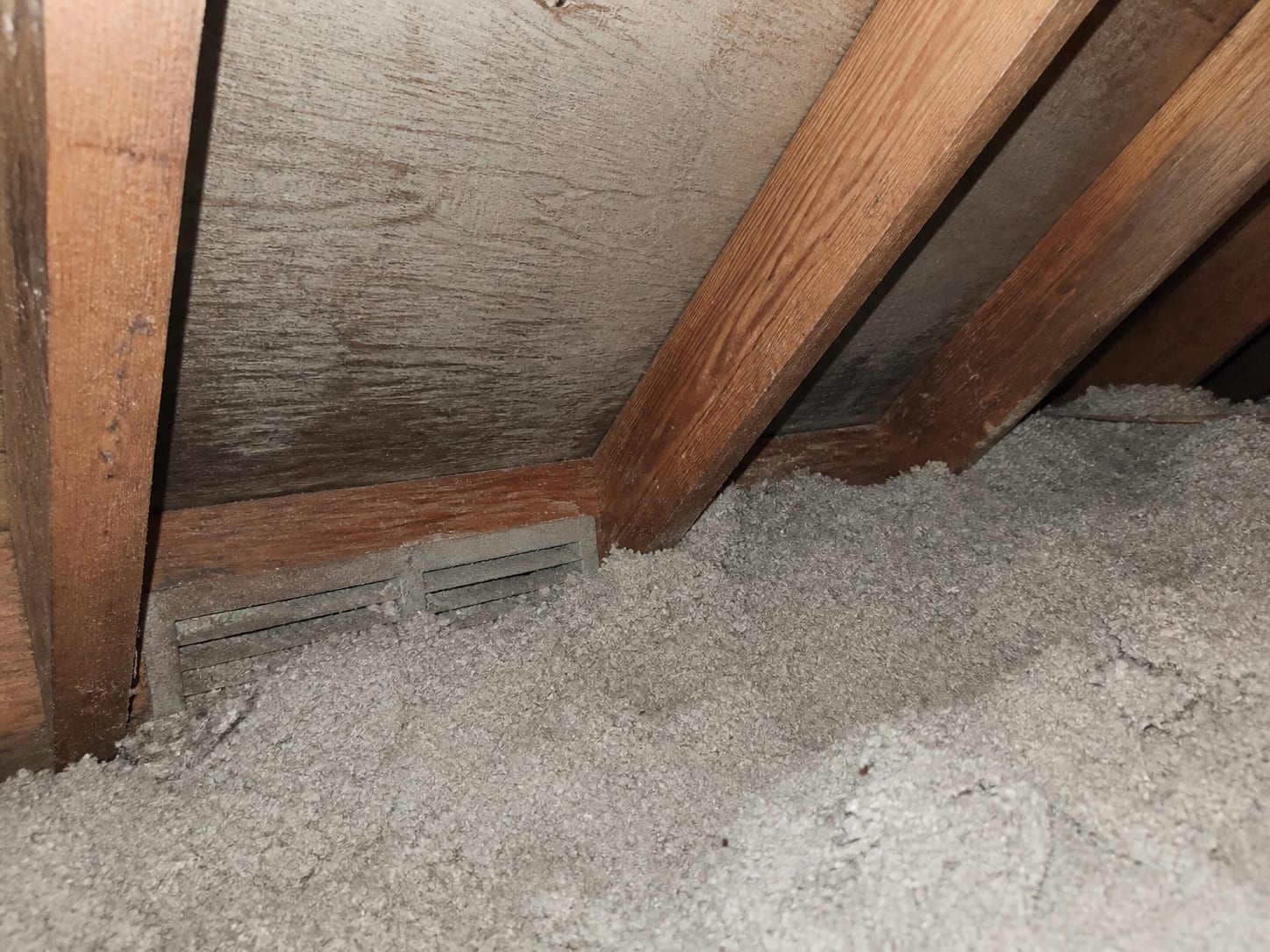 A soffit vent at the eave completely packed with blown insulation pushed hard against it, blocking the airway, viewed from inside the attic near the roof edge, in Plainsboro, New Jersey