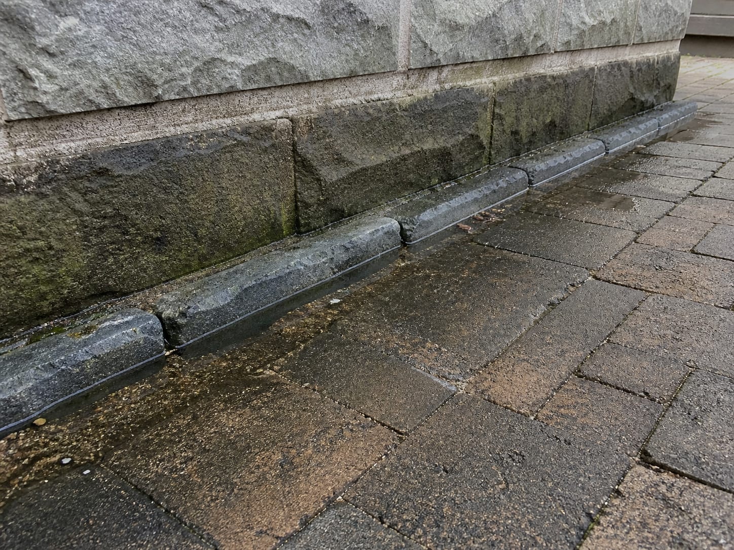 A paver motor court edged in belgian block whose fall runs back toward a masonry foundation, with standing water lying in the joint line and the base of the wall darkened, in Alpine, New Jersey