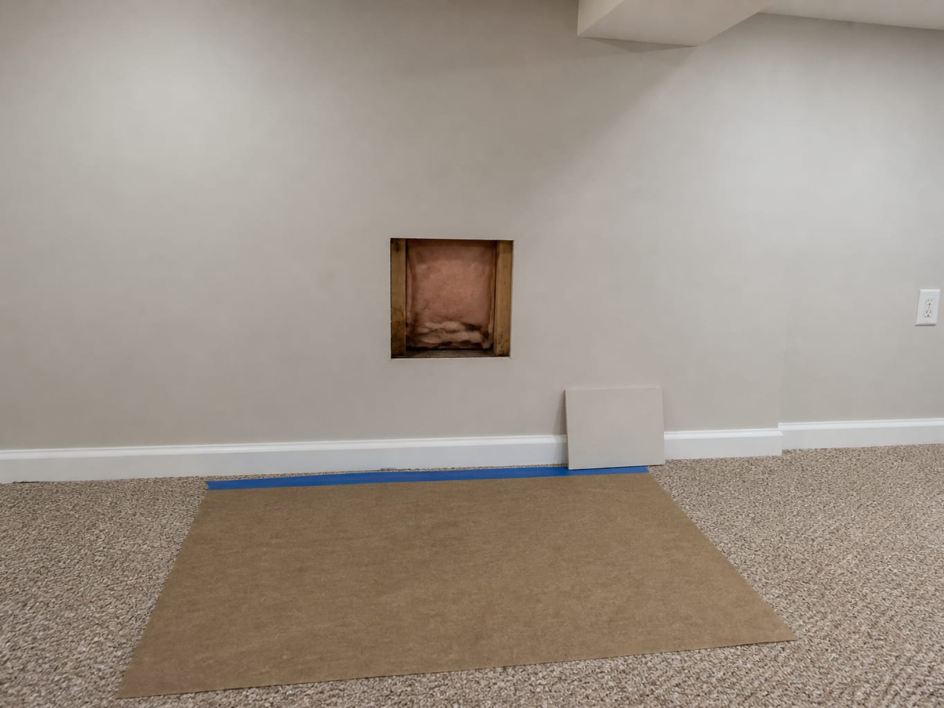 A wider view of a finished basement wall with a small square inspection opening cut between two studs, revealing darkened insulation and framing inside while the rest of the wall stays intact