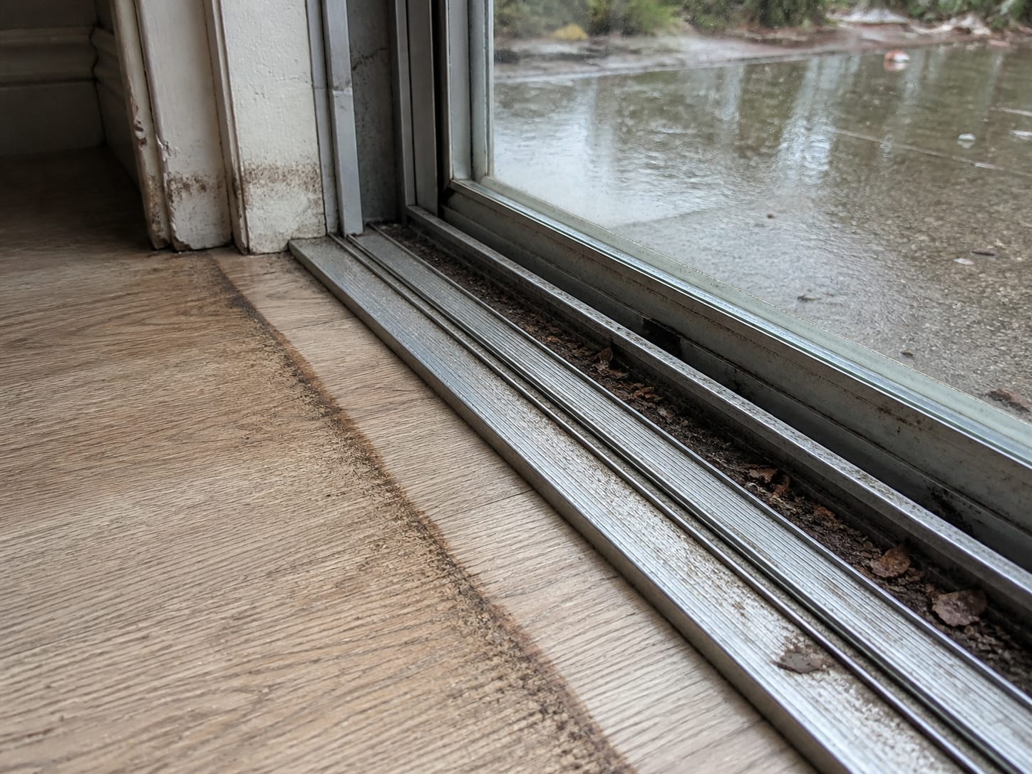 A low close view from inside at floor level along the aluminium track of a sliding patio door at grade, wet leaf debris packed into the channel, a dark tide line running across the flooring at the track and up the bottom of the jamb, the exterior patio slab beyond sitting level with the floor, in Plainsboro, New Jersey