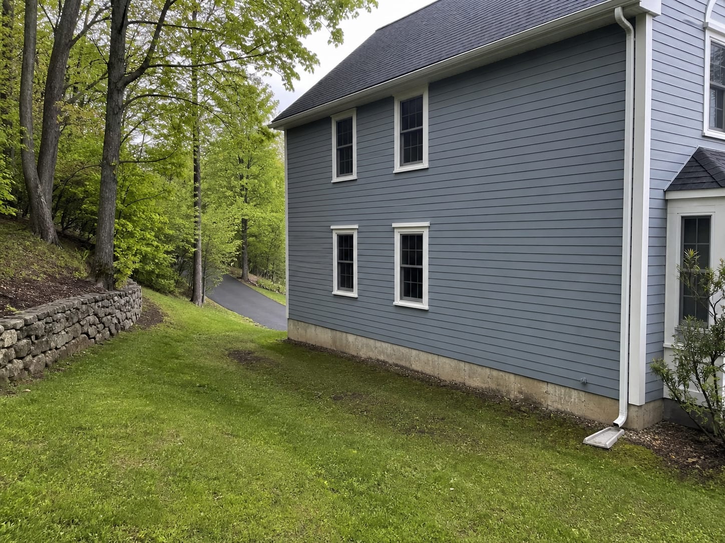 A lawn falling toward the foundation of a clapboard colonial with a downspout discharging beside the wall on a sloping Warren, New Jersey lot