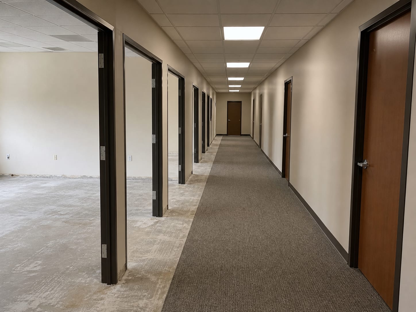 A corridor of a nonresidential building with the doors along one side standing open onto emptied rooms, floor coverings removed to bare slab, and the corridor itself still finished and in use, in Alpine, New Jersey