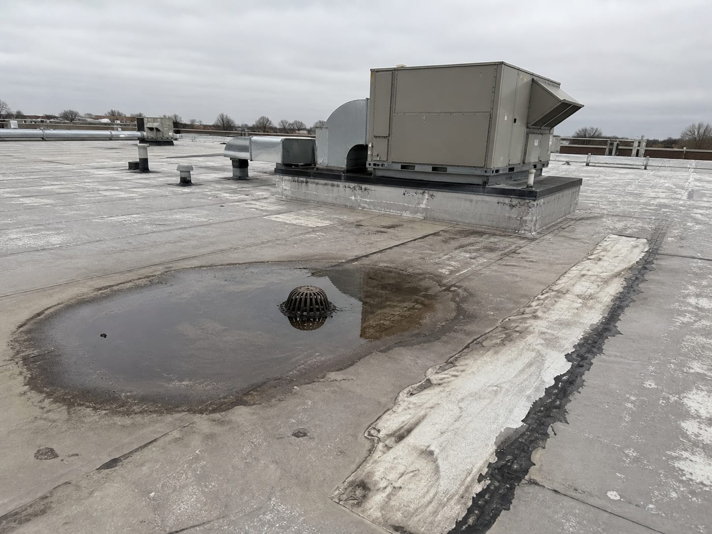 A wide view across a flat low rise commercial roof where water stands around a roof drain beside a patched membrane seam, with a rooftop equipment curb behind it under flat overcast light