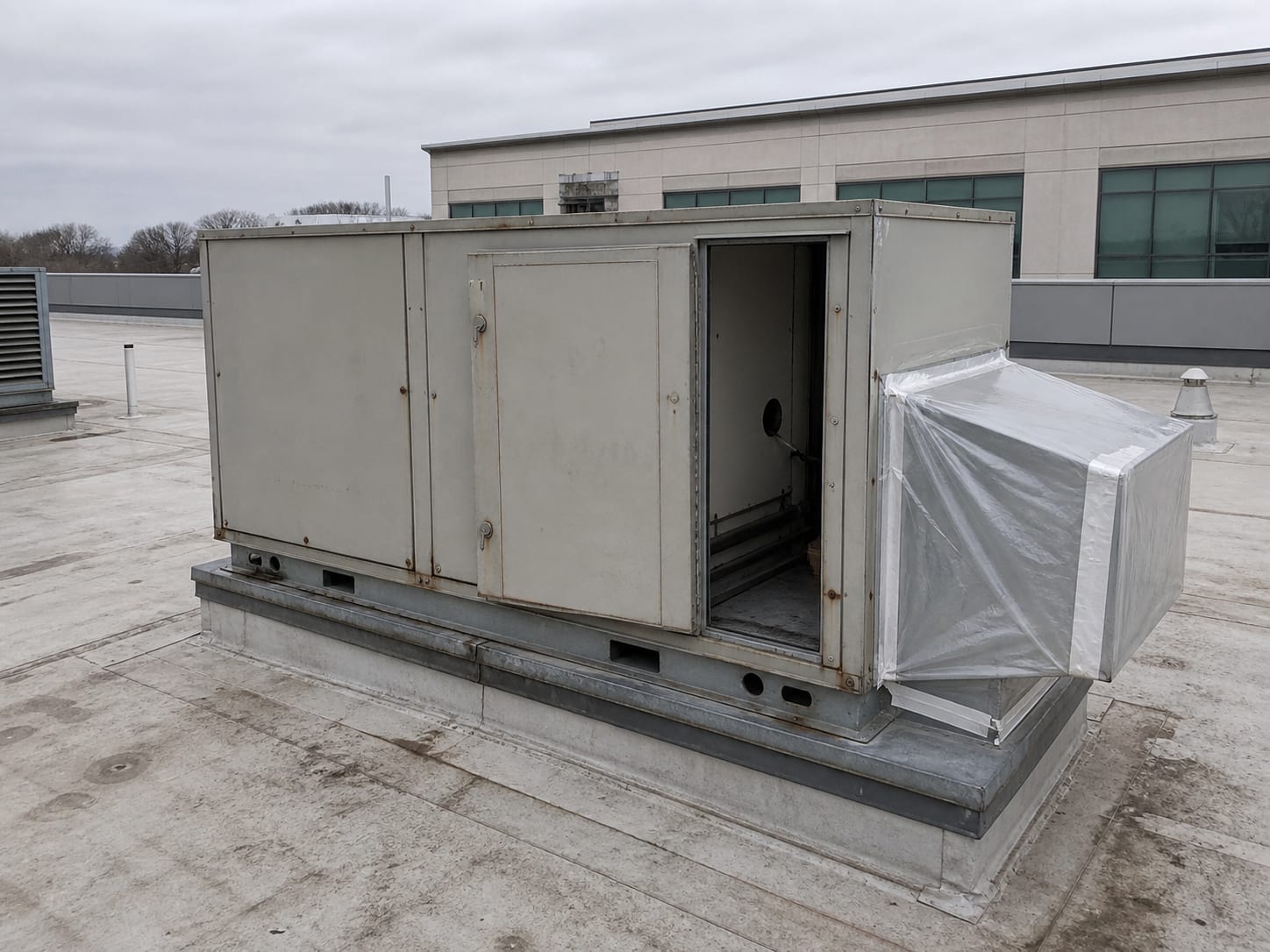 A rooftop air handling unit with a service panel open and temporary poly sheeting sealing one duct connection, overcast flat daylight, no people, in Plainsboro, New Jersey