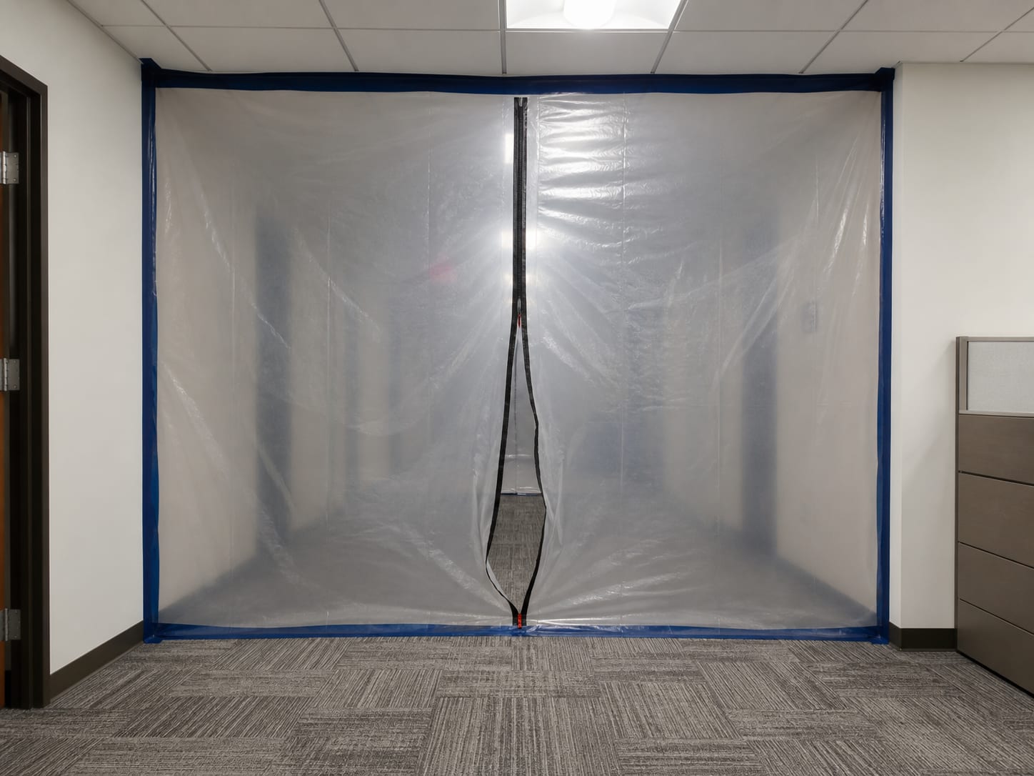 A sealed containment barrier of translucent polythene sheeting spanning a commercial corridor in a West Windsor, New Jersey office building, with a zippered access panel down the centre