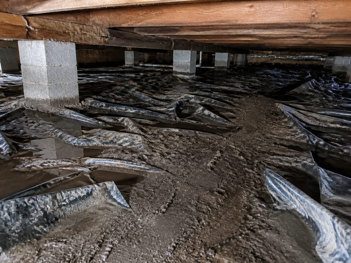 Bare damp soil across a crawl space floor with a torn polyethylene vapour barrier pushed aside and rucked up, shallow water standing in the low spots, seen at a low angle between the piers, in Plainsboro, New Jersey