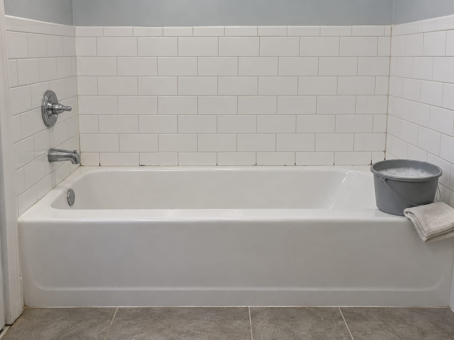 A tiled bathroom wall in a New Jersey home with grout lines, a plain bucket of soapy water and a folded cloth on the edge of the tub