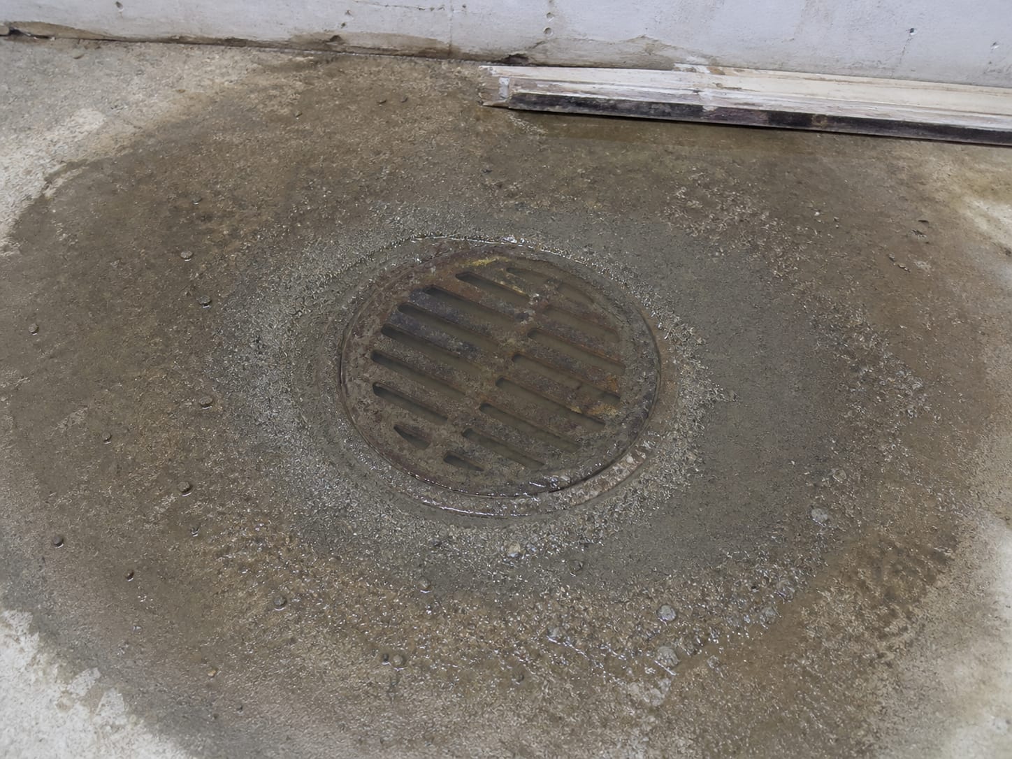 A cast metal floor drain set in a bare concrete slab with water standing above the grate and a ring of fine silt settled around it, in Alpine, New Jersey