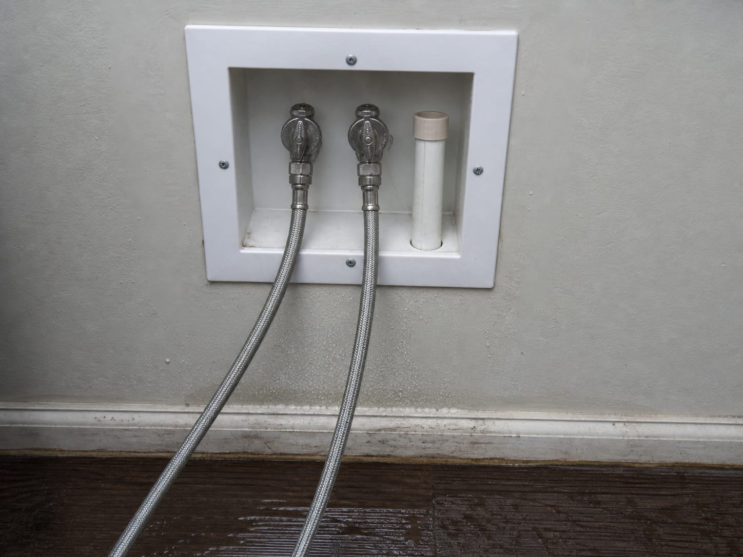 A close view of the hot and cold supply valves at a recessed laundry connection box turned to the closed position, with braided hoses, a drain standpipe and wet flooring below