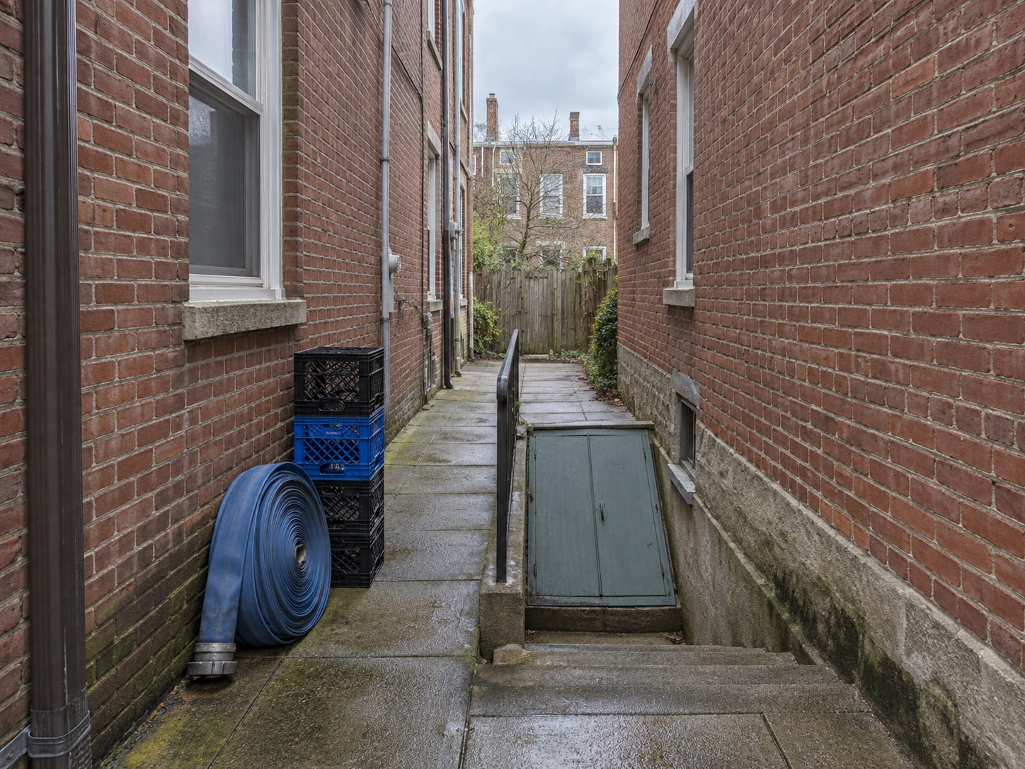 A narrow paved passage between attached brick rowhouses with a coiled hose and steps down to a cellar door in Trenton, New Jersey