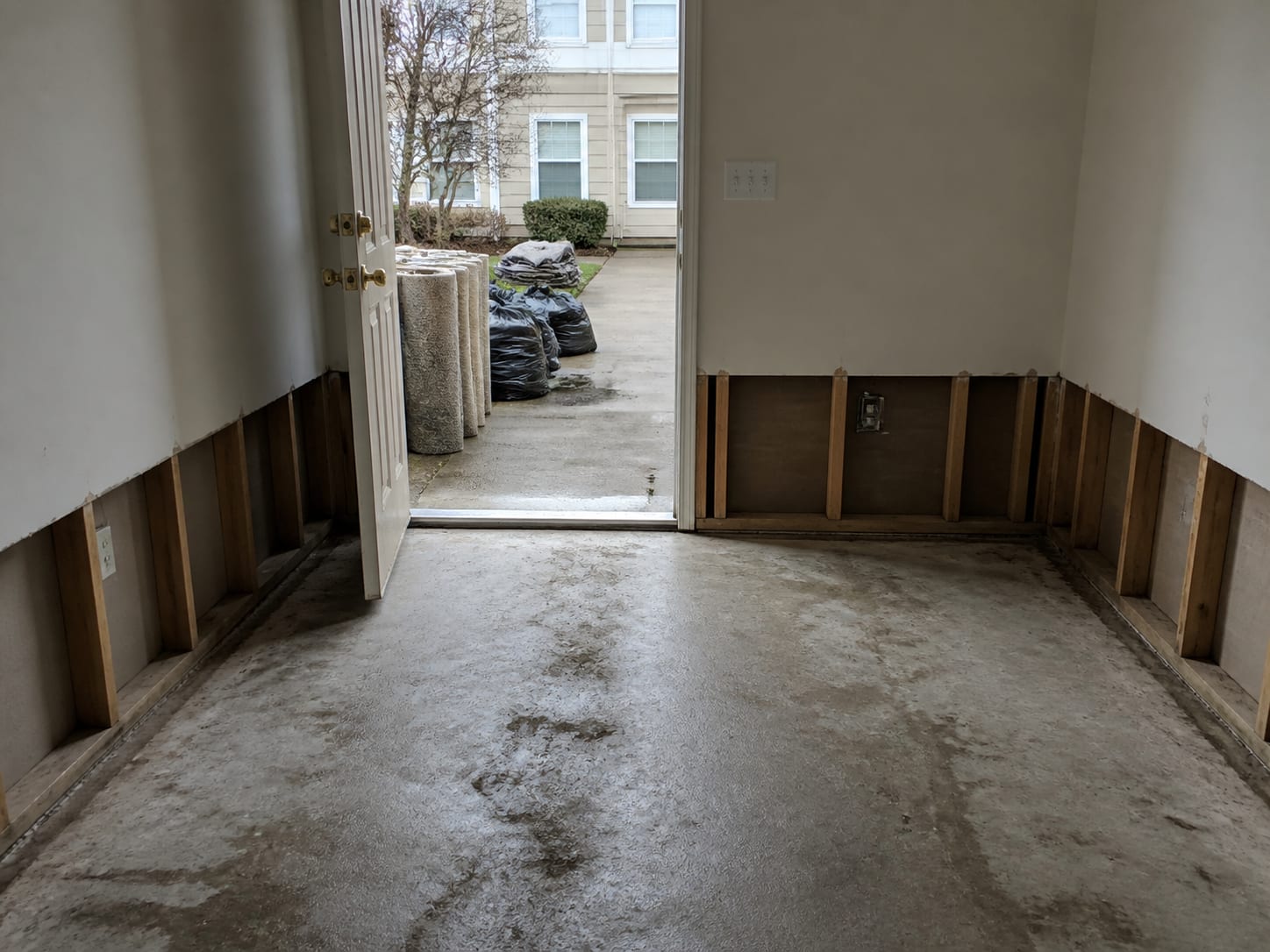 A wider view through an open exterior doorway where rolled flood soaked carpet and sealed disposal bags are staged outside on the walkway, the room behind stripped to bare slab
