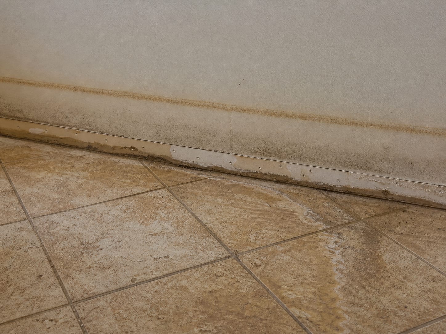 A brown floodwater silt line across a tiled floor and up the base of the drywall in a West Windsor, New Jersey home, with the skirting board removed along one section