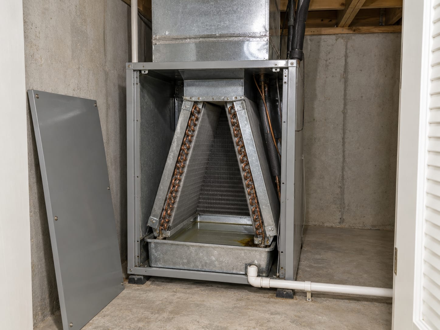 A residential air handler in a New Jersey utility closet with its access panel removed, showing the cooling coil, the drain pan beneath it and a white PVC condensate line