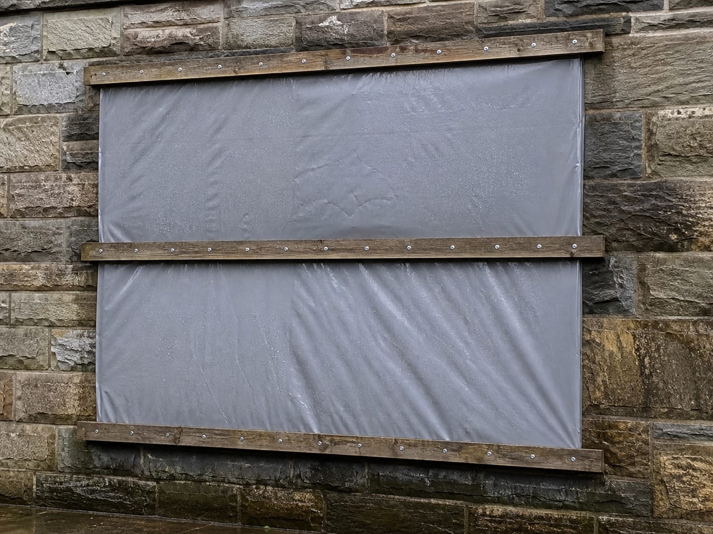 A taut tarpaulin lashed over an exterior opening and held with timber battens screwed to the wall, the masonry around it rain darkened, in Alpine, New Jersey