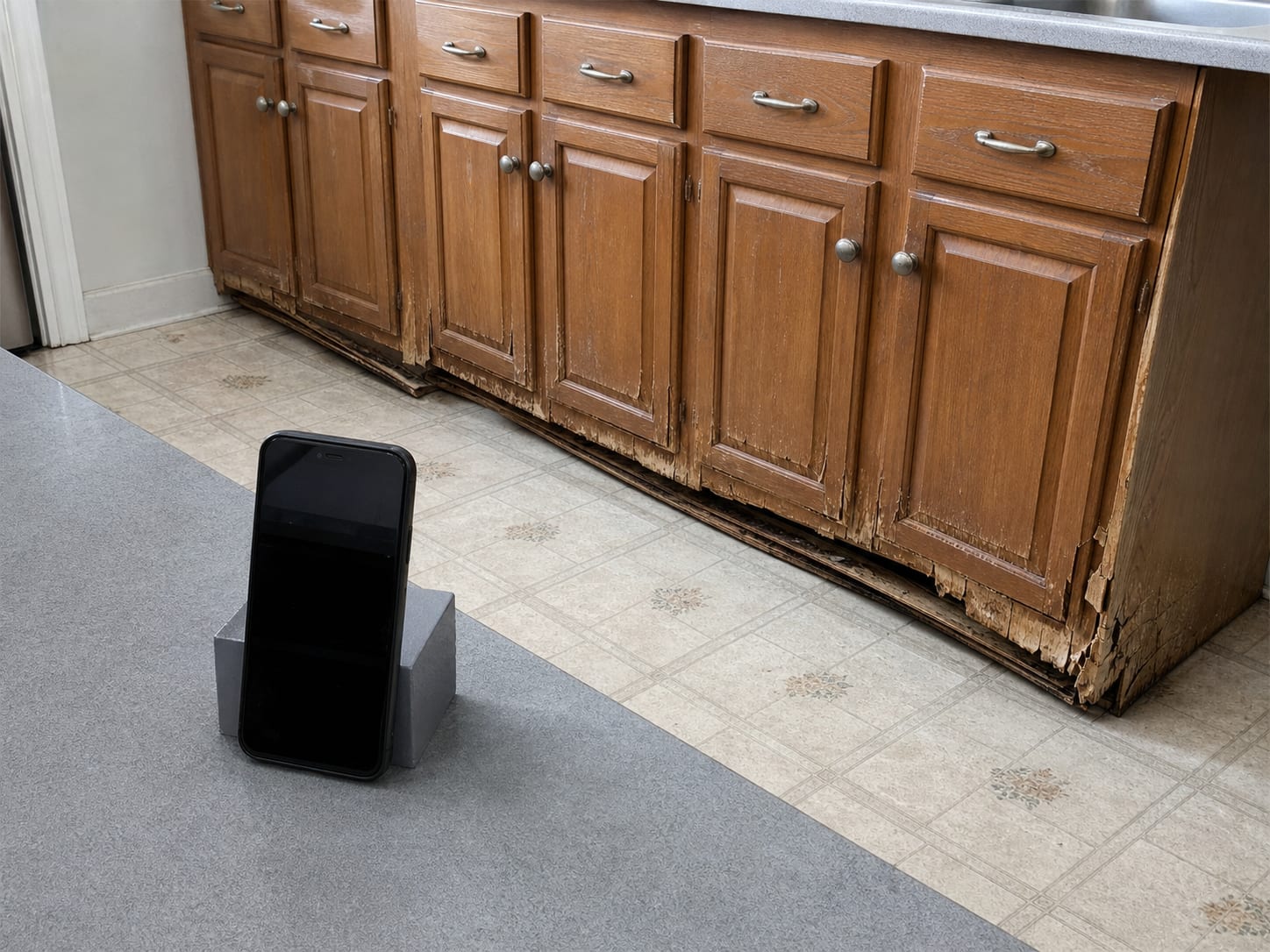 A phone propped upright on a kitchen counter framing a water damaged run of base cabinets, the swollen cabinet ends and the lifting toe kick clearly in view, nothing else on the counter, in Plainsboro, New Jersey