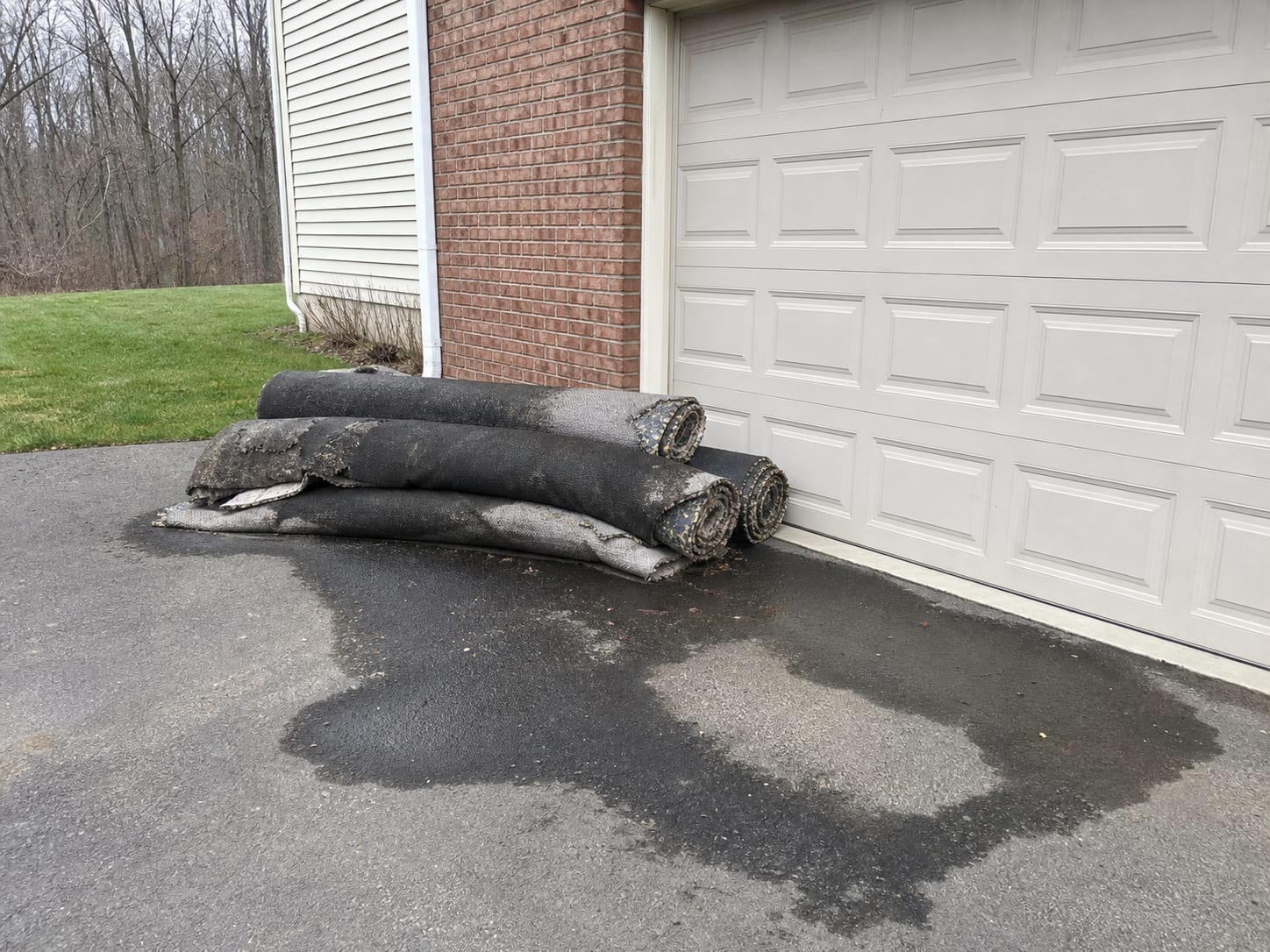 Rolled sections of removed wet carpet and underlay stacked on a driveway beside a closed garage door, dark with water and left in place, overcast flat daylight, in Plainsboro, New Jersey
