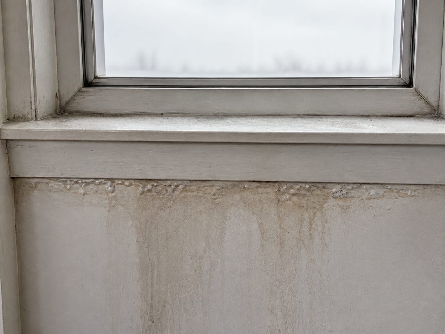 A close view of the underside of a window stool and the wall beneath it, showing water staining, lifted paint and darkened joints at the frame