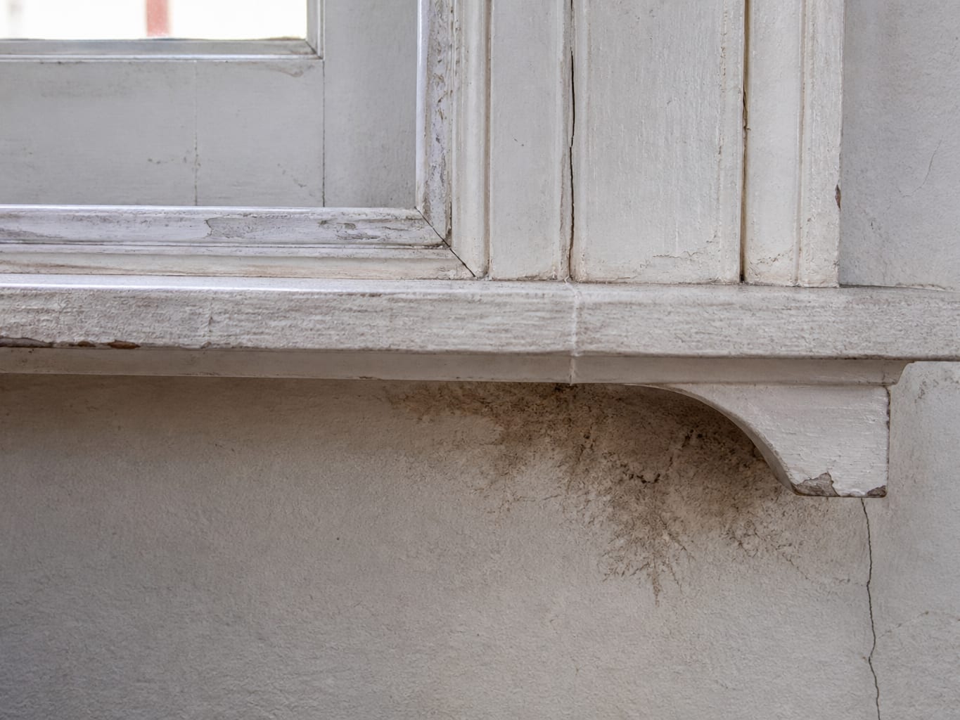 A close view of dark staining and blistered paint on the wall below a mid-century wood window stool in a Livingston home, evidence of water entering at the window
