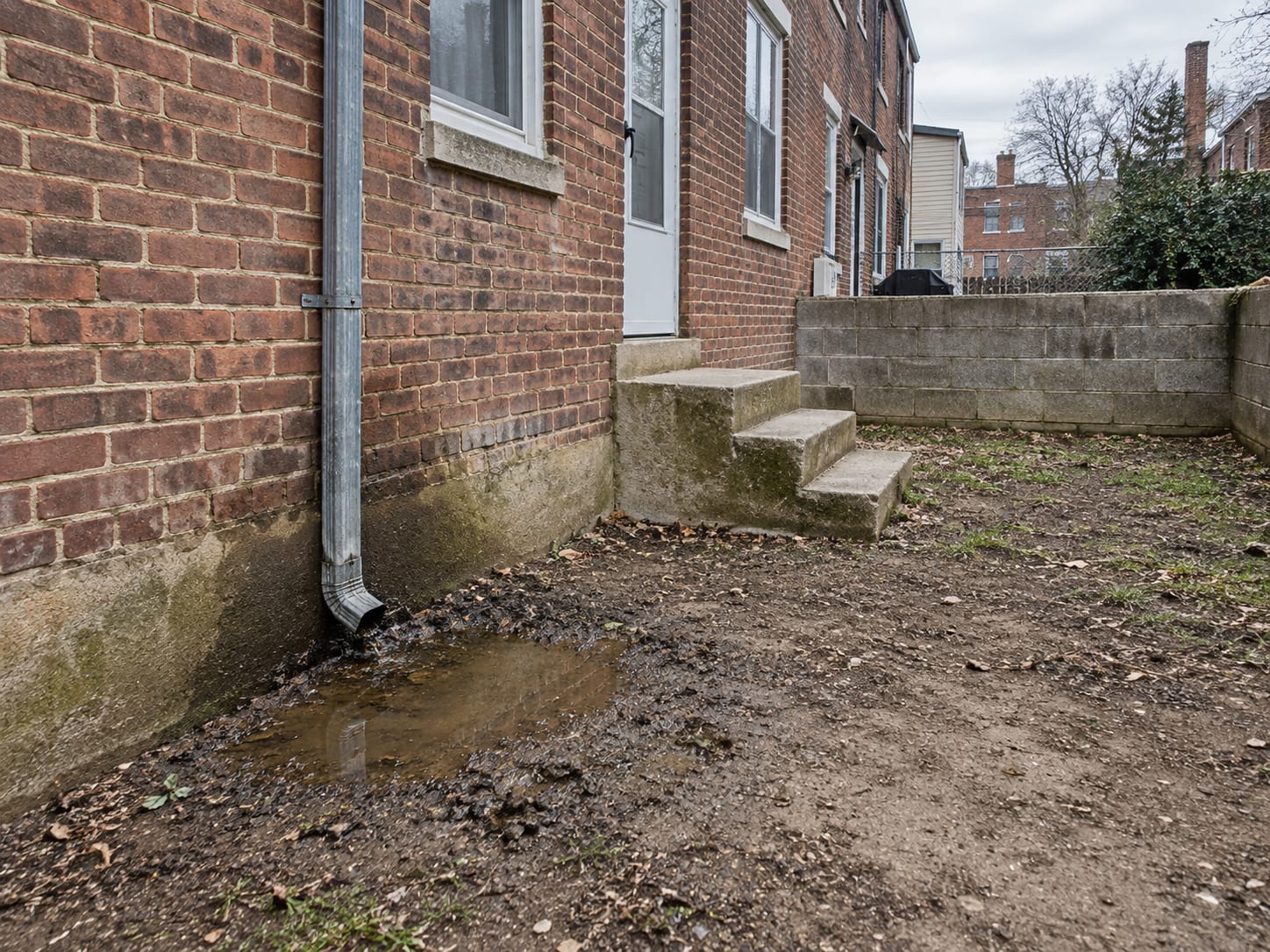 Rear yard of a Trenton, New Jersey rowhouse where a downspout ends against the brick foundation and water has ponded on the packed soil