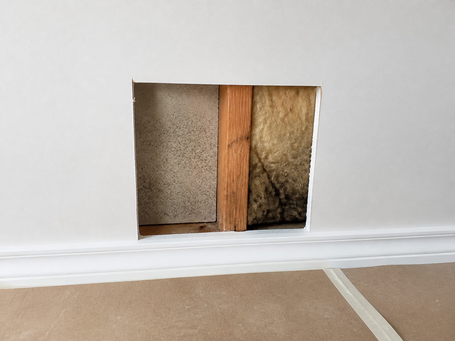 A small inspection opening cut in drywall exposing a stud bay, the back of the board and damp fibreglass insulation, in Warren, New Jersey