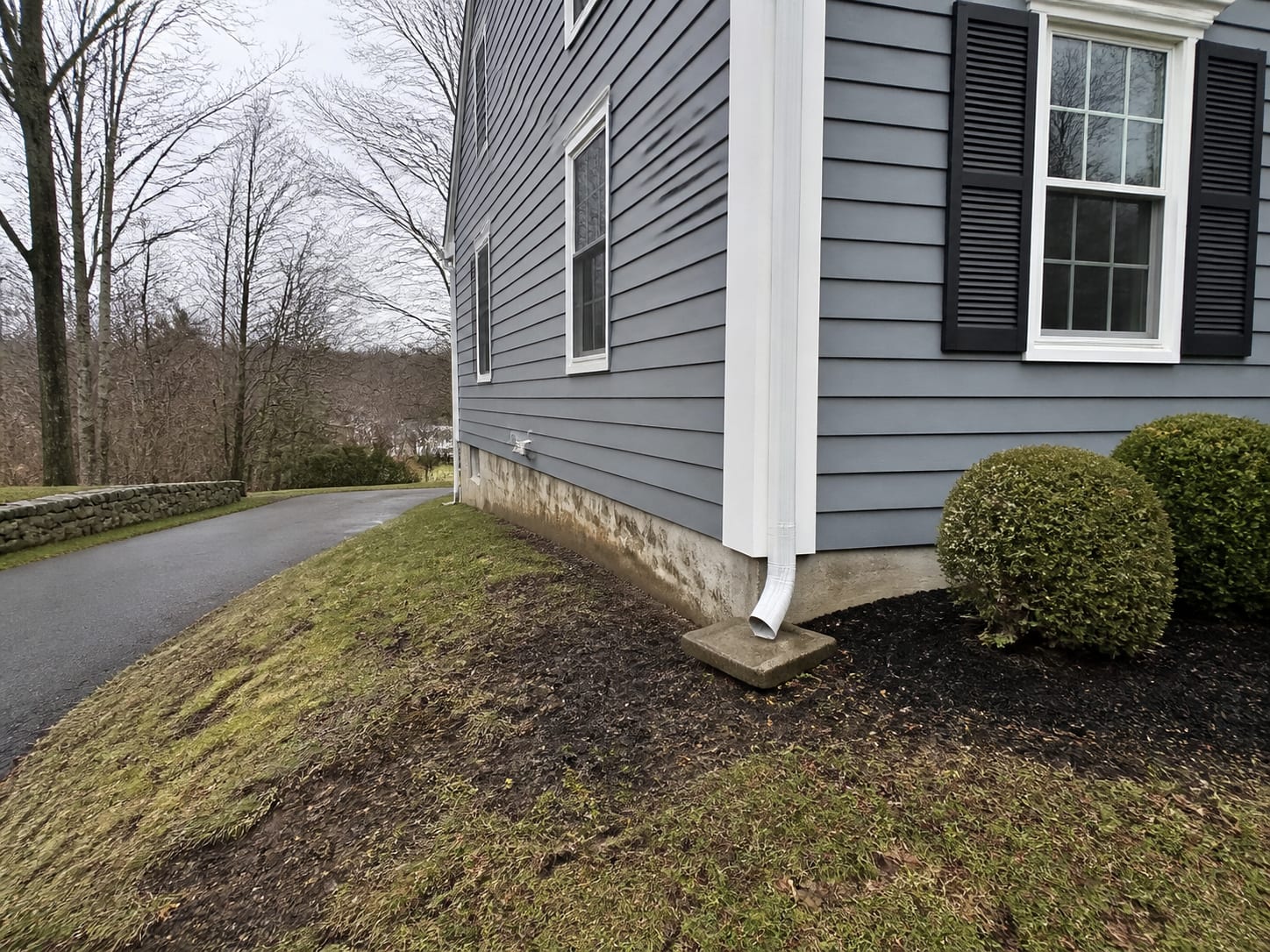 A downspout discharging directly beside a foundation on a lawn that falls toward the house, beside a painted clapboard colonial in Warren, New Jersey