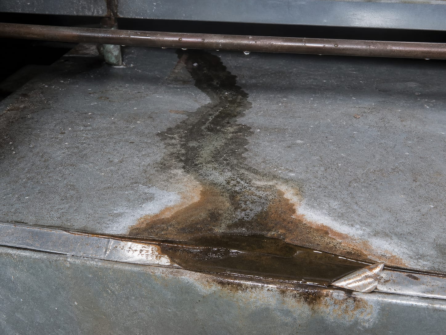 A dark wet trail running across the top of a sheet metal plenum below an air handler, with beaded moisture on the cold line above and corrosion where the water has been sitting, in Alpine, New Jersey