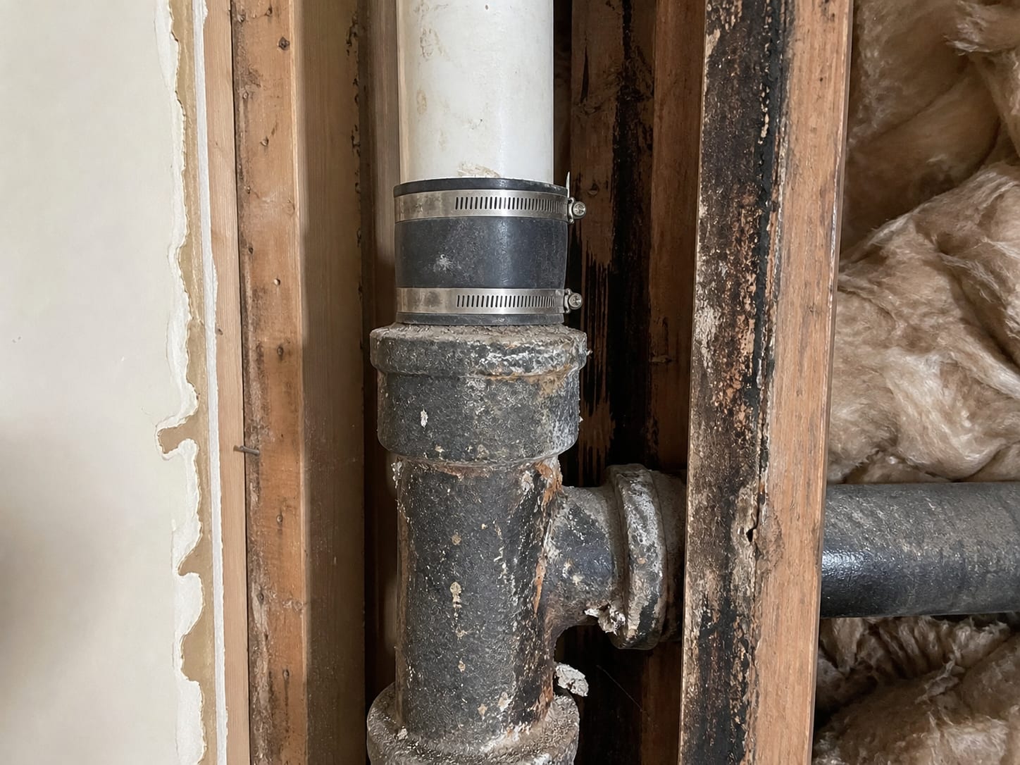 A close view of a shared vertical drain and vent stack inside an opened wall chase, with a hub joint above and dark staining tracking down the framing beside it