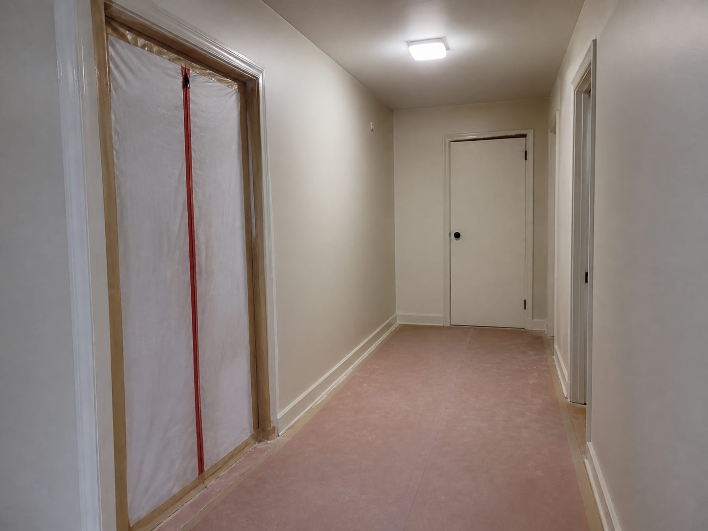 A wider view of a townhouse hallway with containment sheeting and a zipper closure sealing one doorway while the adjacent doorway is closed and untouched, floor protection running between them