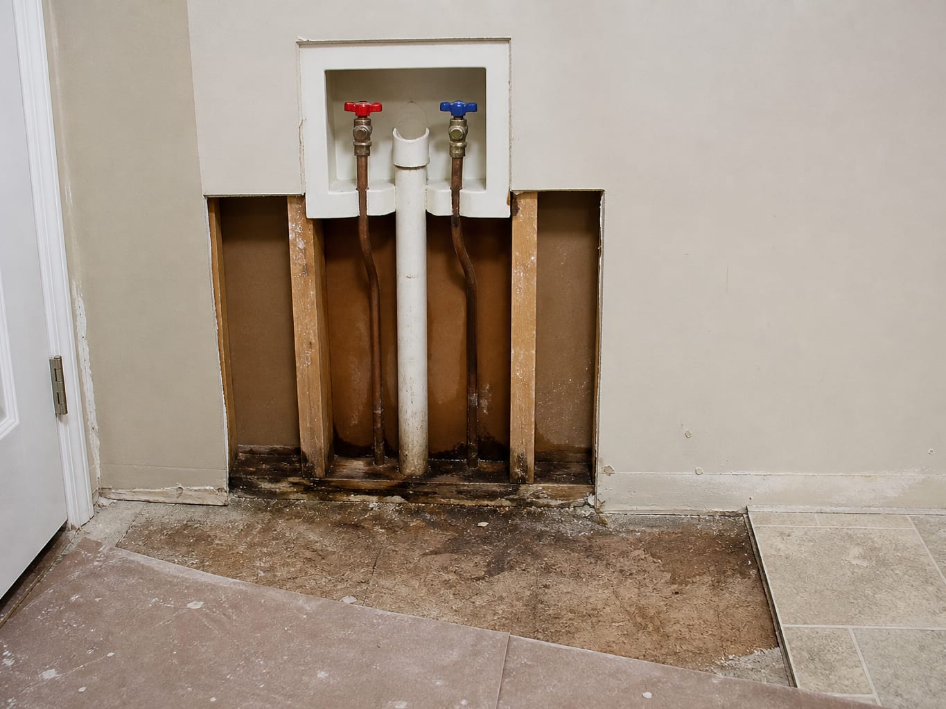 A wider view of a laundry alcove where the wall around the recessed supply valve box has been opened, exposing the shutoff valves, the supply lines and the darkened framing behind them