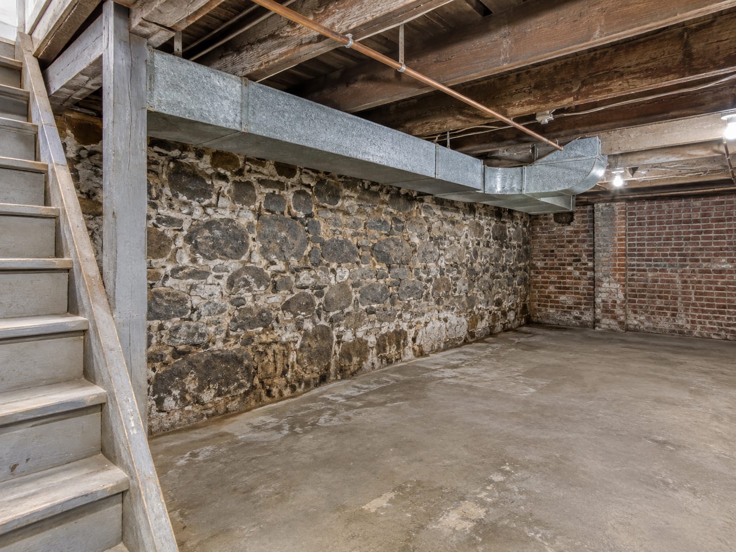 Rowhouse cellar in Trenton, New Jersey with a fieldstone foundation wall, exposed floor joists and a run of ductwork overhead