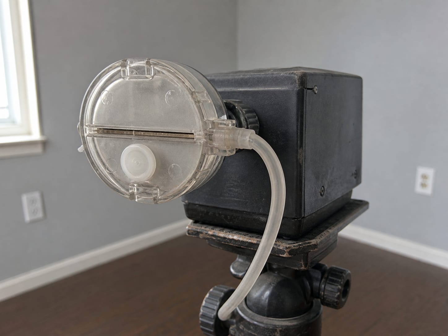 A close view of a sealed spore trap cassette seated on the inlet of an air sampling pump on a tripod, the cassette body and its collection slot sharply detailed, the room soft behind it, in Plainsboro, New Jersey