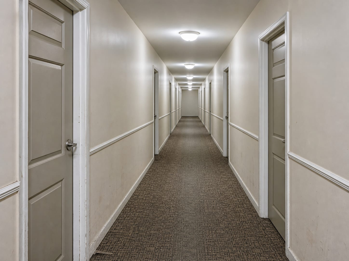 A condominium common corridor seen down its length, closed painted panelled unit doors at intervals on both sides, painted drywall walls and patterned low pile carpet, no people, in Plainsboro, New Jersey
