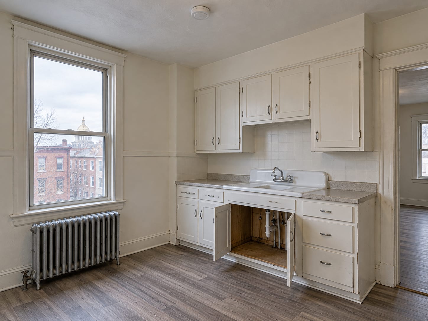 Empty rental apartment kitchen in Trenton, New Jersey with a window, a radiator and the cabinet under the sink standing open