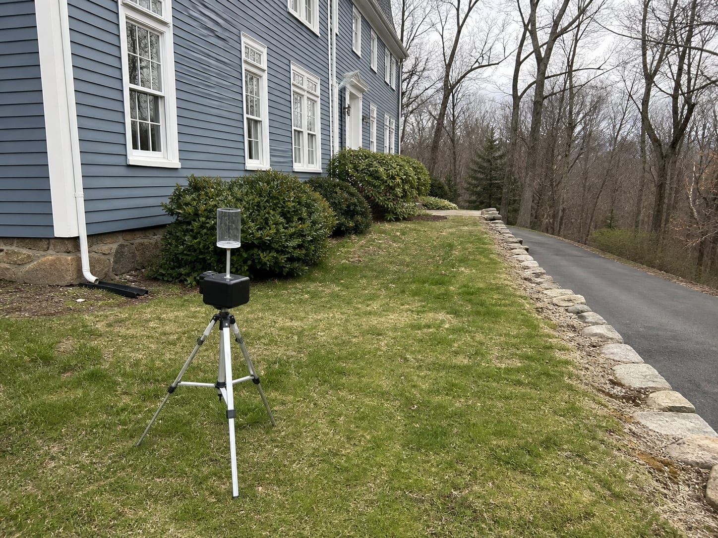 An air sampling pump on a low tripod set on a lawn beside the foundation of a painted clapboard colonial, taking the outdoor reference sample, in Warren, New Jersey