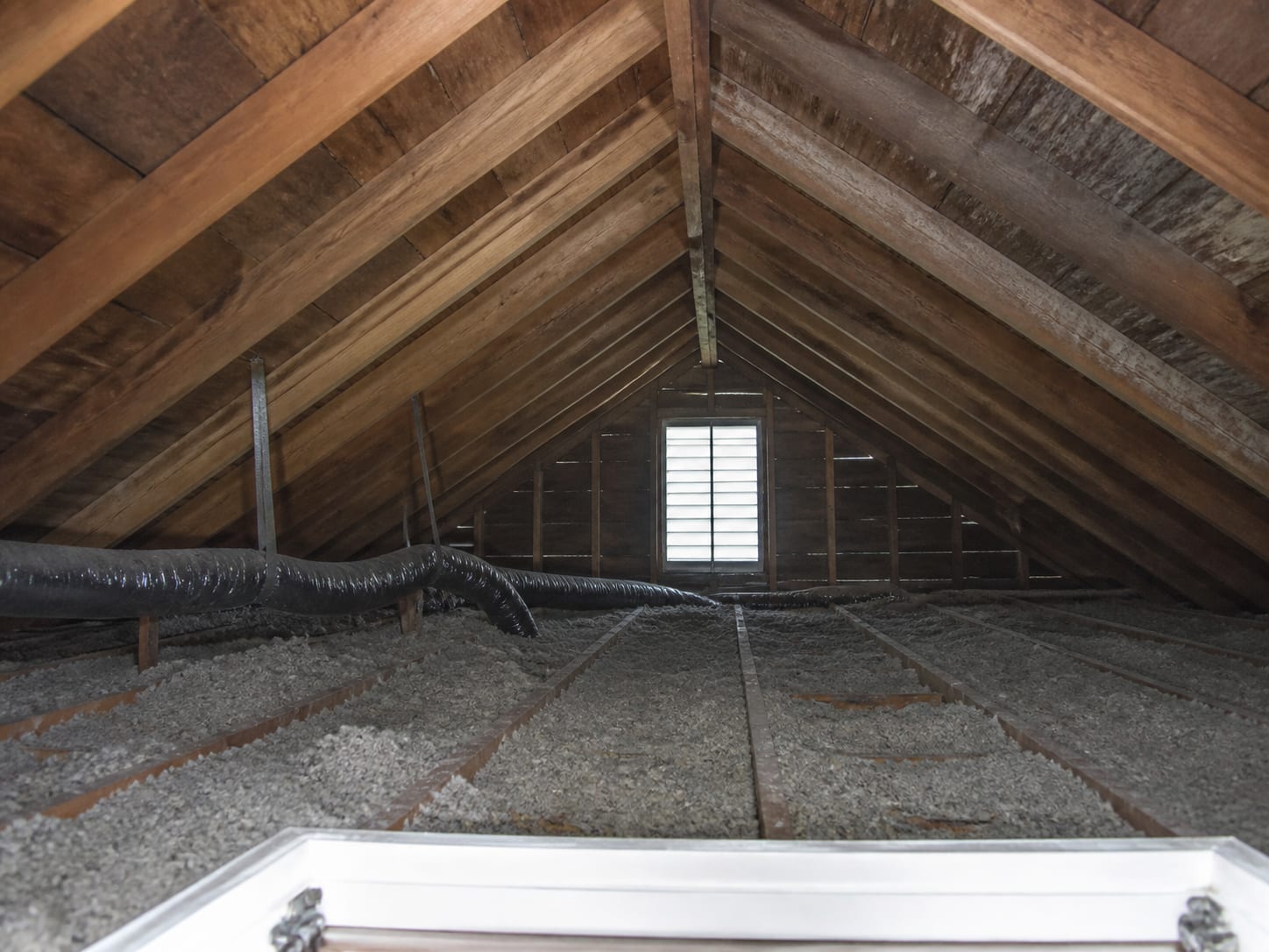 An opened attic hatch in an older West Windsor, New Jersey house showing rafters, a gable vent admitting daylight and blown insulation across the ceiling joists
