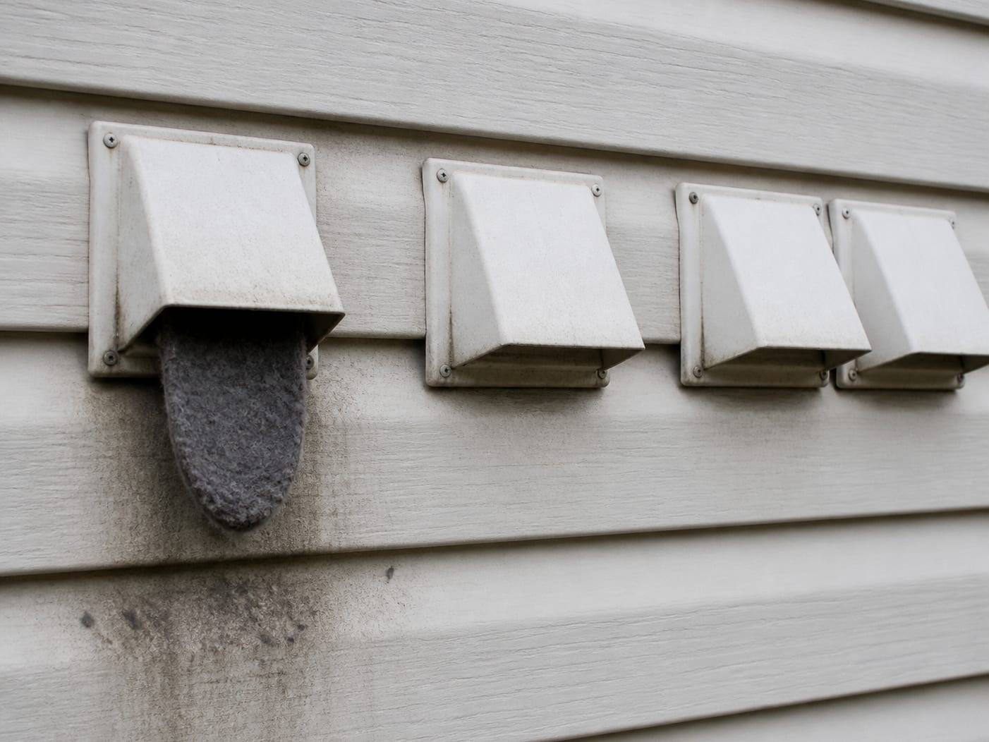 A bank of dryer exhaust wall caps on the exterior of a New Jersey apartment building with a mat of grey lint caught at the damper of one of them