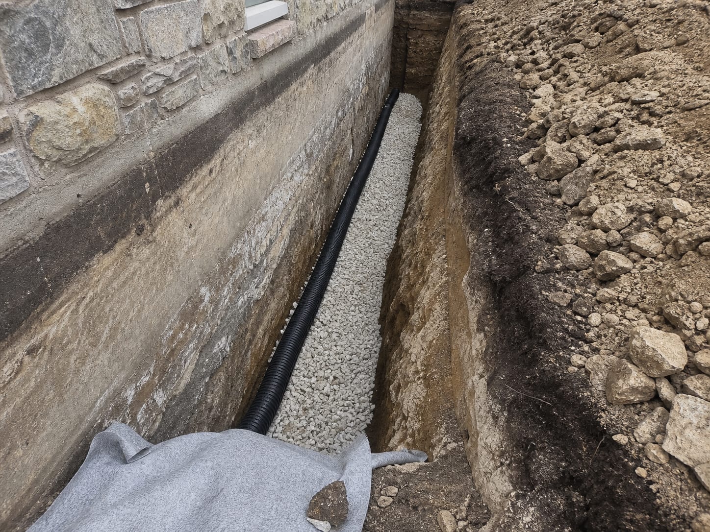 A close view into an open trench where perforated pipe sits in clean washed gravel against a foundation wall, with filter fabric folded back and the wall face brushed clean, in Alpine, New Jersey