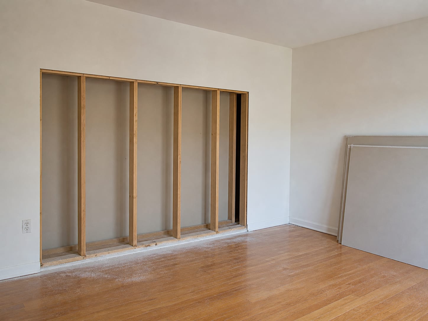 An opened and dried wall bay with clean pale framing exposed and the drywall cut edge running straight and level above it, new plasterboard sheets leaning unused against the opposite wall of the room, in Plainsboro, New Jersey