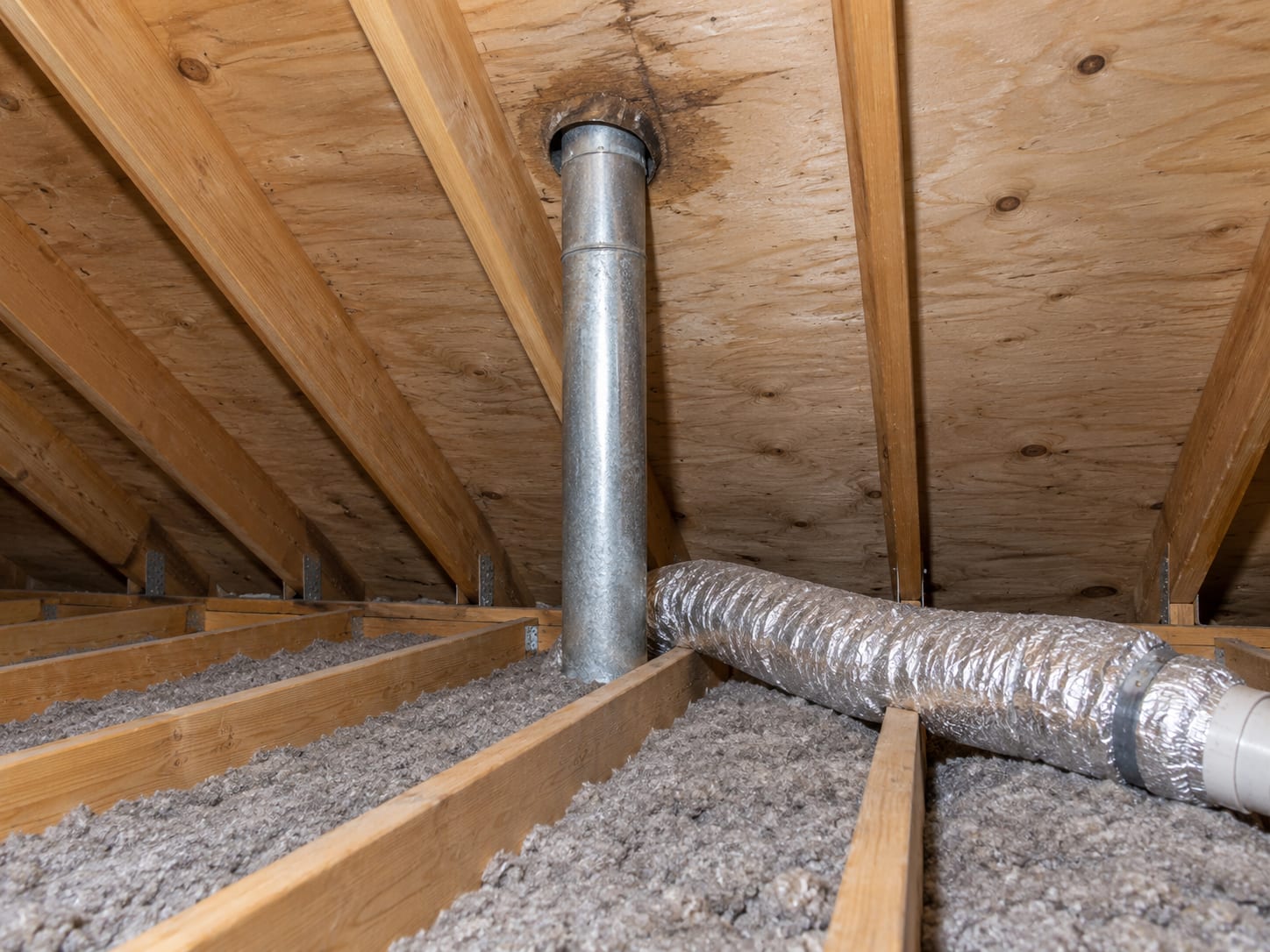 The underside of a pitched roof in a New Jersey attic with a plumbing vent pipe passing through the sheathing, a bathroom exhaust duct and light staining around the penetration