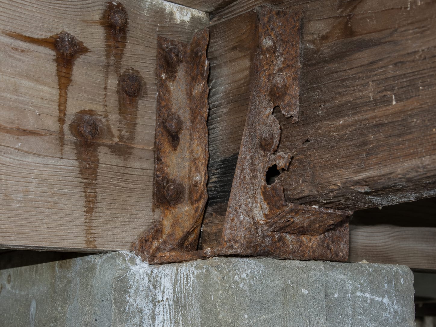 A rusted metal joist hanger at a bearing point with rust staining bleeding into the timber around it and the fastener heads swollen and pitted, in Alpine, New Jersey