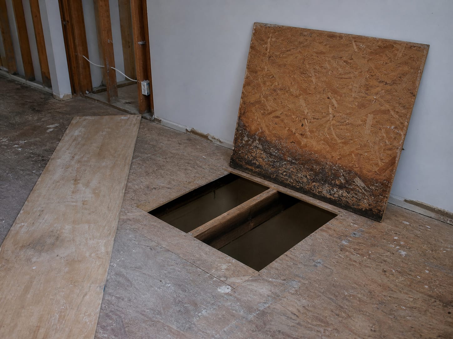 A wider view of a townhome floor in East Windsor where a rectangular section of subfloor sheathing has been cut out between the joists, exposing the framing below and the darkened underside of the cut panel edge