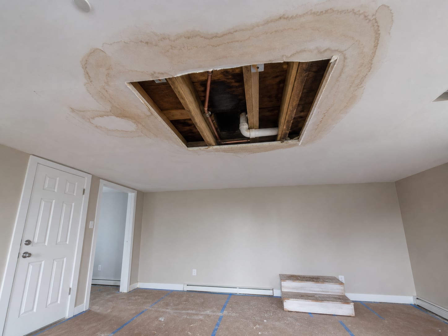 A wider view of a lower unit ceiling opened in a square cut, exposing the joist bay and the supply line that runs to the dwelling above, staining spreading across the drywall around the opening