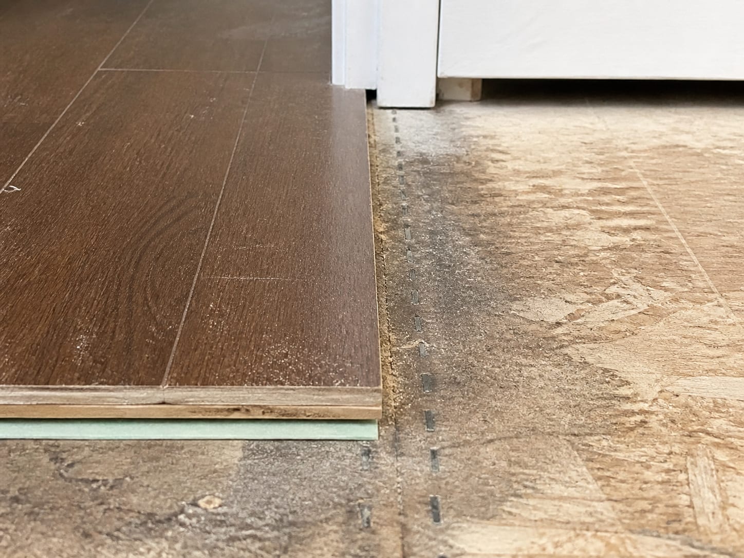 A close view of a doorway threshold where flooring was cut back, showing the layered edge of plank and underlayment beside the bare subfloor where the removed material ended